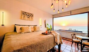 A bedroom with a large bed, a nightstand, a wall-mounted light, a decorative wall piece, and a view of the ocean at sunset.