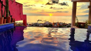 Une piscine à débordement offrant une vue imprenable sur le coucher de soleil sur la mer, avec des palmiers et des bâtiments en arrière-plan.