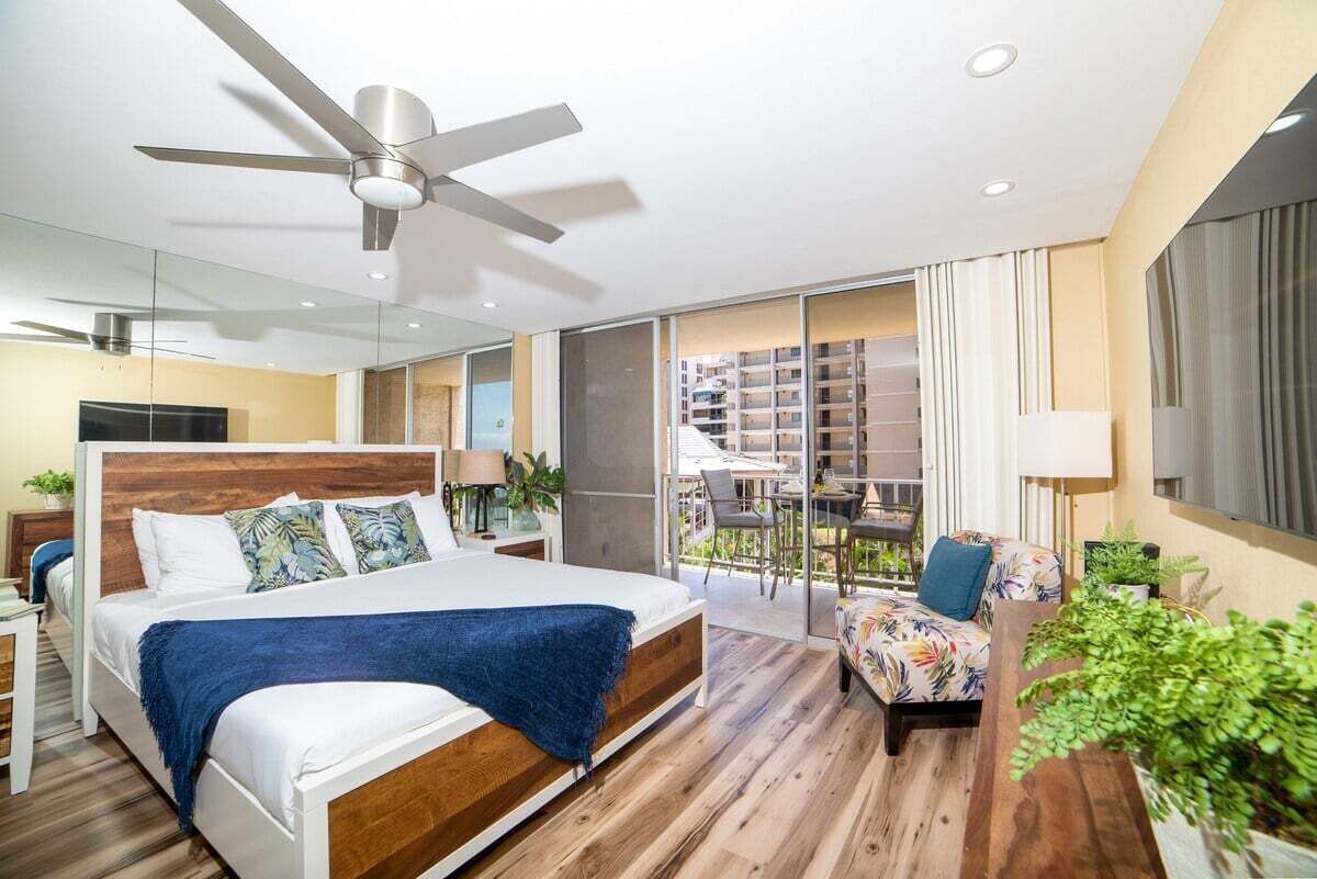 Ein modernes Schlafzimmer mit einem großen Bett, einem Deckenventilator, einem Balkon mit Blick auf ein Gebäude und einer Sitzecke mit einer Pflanze.