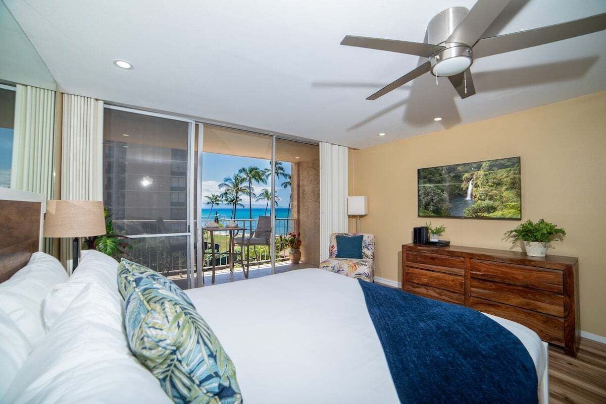 Una camera d'albergo con un letto grande, una ventola a soffitto, un balcone con vista sull'oceano e un comò in legno.