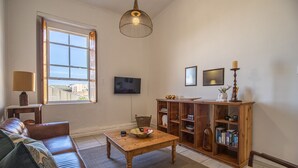 Uma sala de estar com sofá de couro, mesa de centro de madeira, televisão, estante de livros e janela com vista para edifícios.