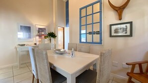 Uma área de refeições com mesa branca, cadeiras de vime, uma tigela e uma garrafa.