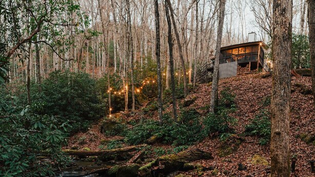 Una cabana al bosc amb un rierol, envoltada d'arbres i fulles d'octubre.