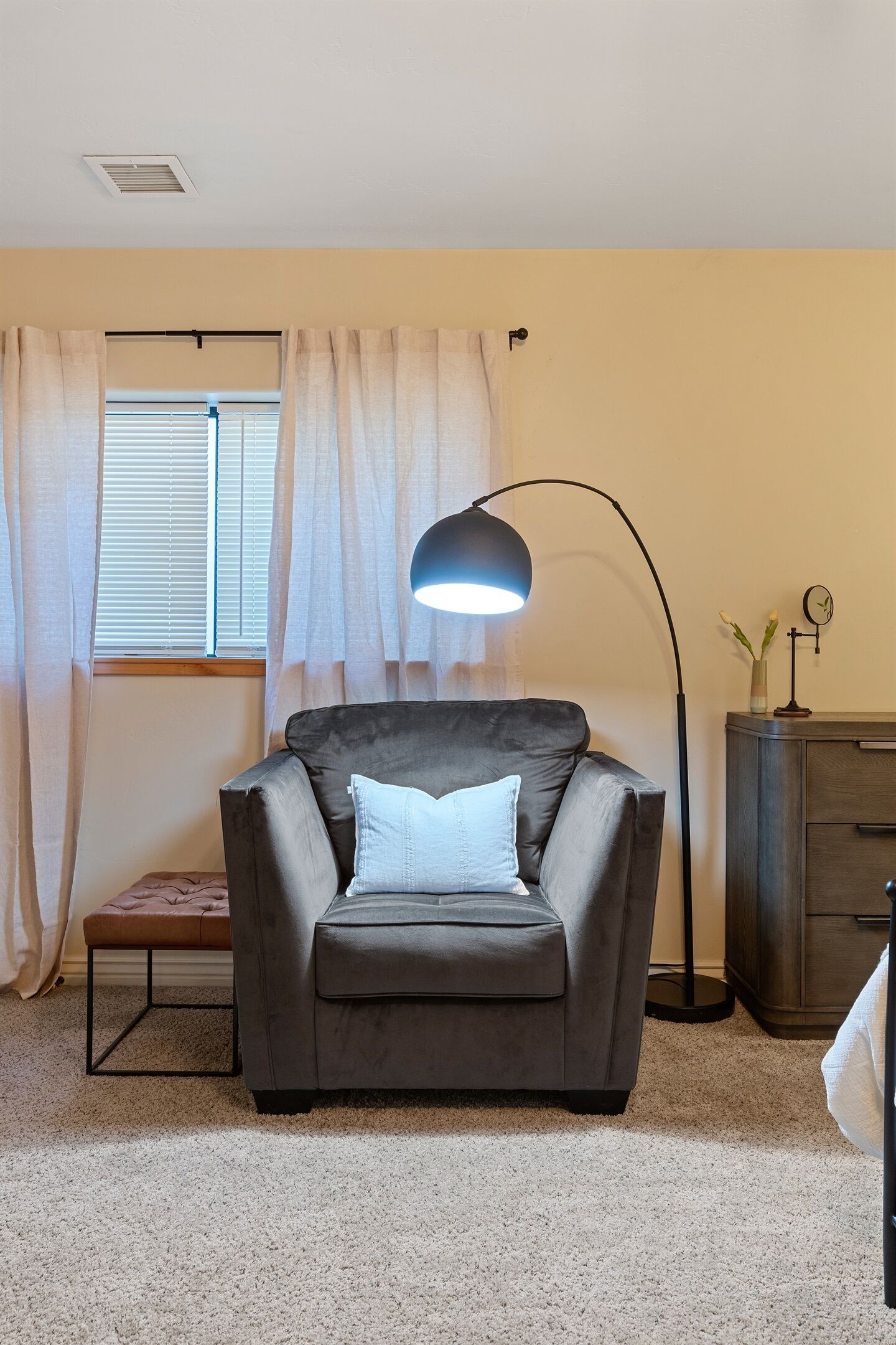 A living room with a dark grey sofa, a small side table, a floor lamp, a window with blinds, and a beige carpet.