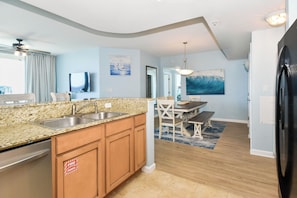A modern kitchen with granite countertops, wooden cabinets, and a built-in dishwasher. There is a dining area with a table and chairs, and a large painting on the wall.