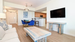 Een moderne woonkamer met een bank, een salontafel en een eethoek met stoelen en een tafel. Er is een keuken met houten kasten en een magnetron. Een flatscreen televisie is aan de muur bevestigd.