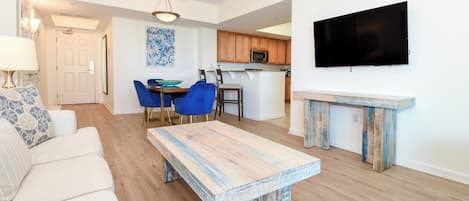 Un salon moderne comprenant un canapé, une table basse et un coin repas avec des chaises et une table. Il y a également un coin cuisine avec des meubles en bois et un four à micro-ondes. Un téléviseur à écran plat est fixé au mur.