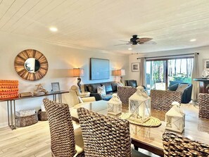 A modern living room with a dining area, featuring wicker furniture, a round mirror, and a ceiling fan.