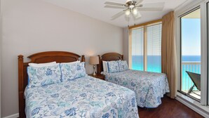 Une chambre avec deux lits, un ventilateur de plafond, une fenêtre donnant sur l’océan et un balcon avec une chaise.
