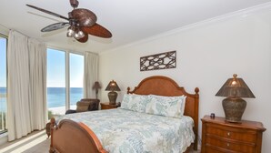 Una camera da letto con un letto grande, un ventilatore a soffitto, un comodino con lampade e vista sull'oceano attraverso ampie finestre.