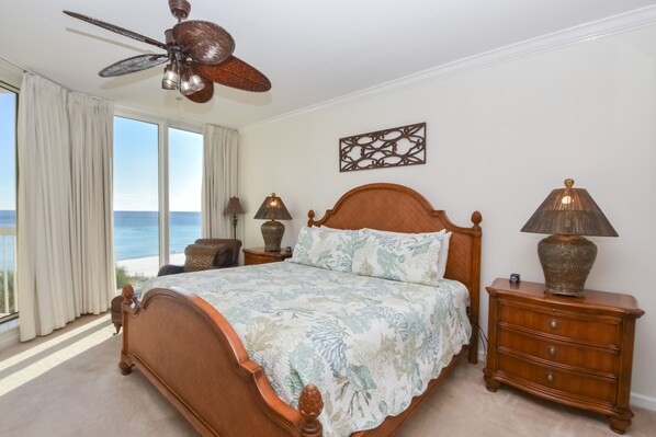 Quarto com uma cama grande, um ventilador de teto, mesa de cabeceira com luminárias e vista para o oceano por grandes janelas.