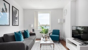 Een moderne woonkamer met een grijze bank, kussens in teal-tint, een salontafel, een blauwe fauteuil, een televisie en een groot raam met uitzicht op gebouwen.