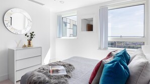 Een moderne slaapkamer met een groot raam, een ronde spiegel, een witte dressoir en een zachte poef.
