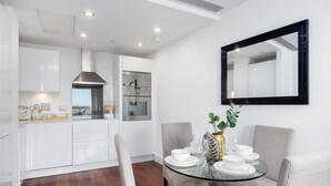 Een moderne keuken met witte kastjes, een roestvrijstalen afzuigkap en een oven. Er is een eethoek met een glazen tafel, witte stoelen en een spiegel aan de muur.