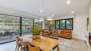 Ampla sala de estar com mesa de jantar de madeira, sofá de couro e grandes janelas com vista para o exterior.