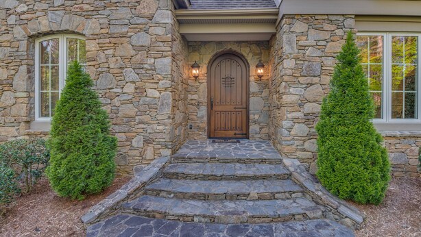 A stone house with a wooden front door, two potted evergreen trees, and a stone staircase leading to the entrance.