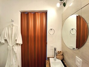 Une salle de bain avec une porte en bois, un peignoir blanc accroché à un crochet, un miroir rond et un lavabo.