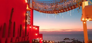 Un hôtel en bord de mer, avec une façade rouge, une pergola recouverte d’un treillis et illuminée de guirlandes lumineuses, et une vue sur la mer au coucher du soleil.