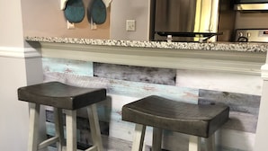 A kitchen with two wooden bar stools, a stainless steel refrigerator, and a decorative wall shelf.