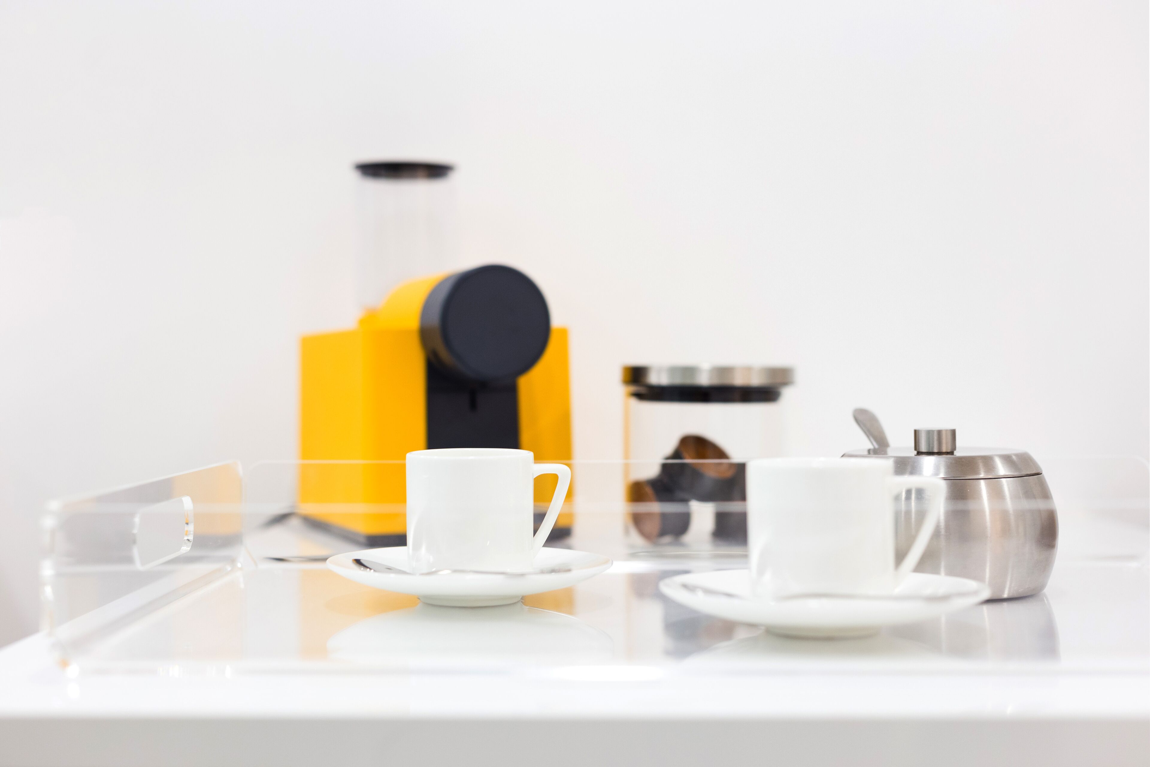 A clear glass table with two white cups and saucers, a yellow coffee machine, and a silver espresso maker.