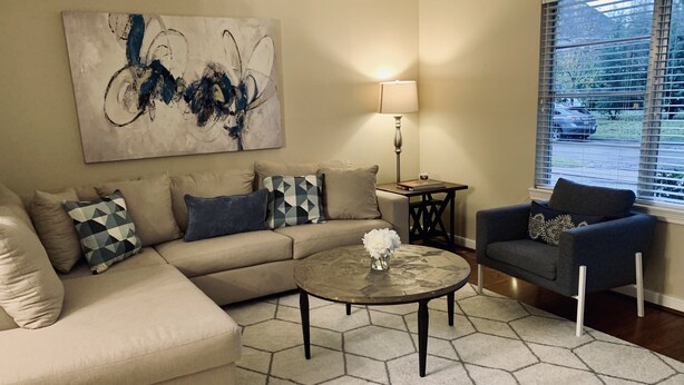 A living room with a beige sectional sofa, a blue armchair, a coffee table with a vase of flowers, a floor lamp, and a painting on the wall. - Inviting/Cozy Home in Medical Center - Beautiful home to stay!!  (Houston)