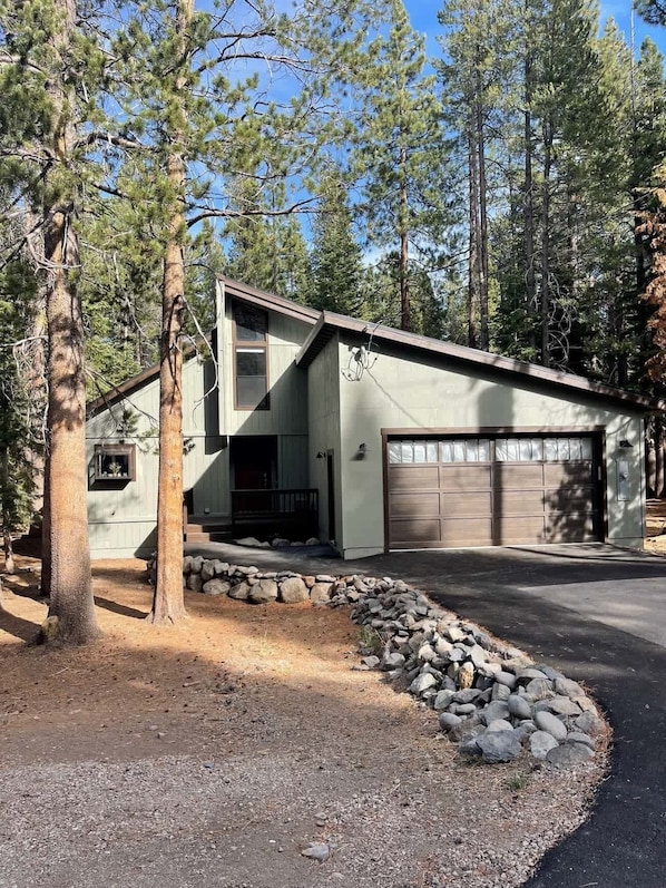 A modern single-story house with a garage, surrounded by a stone retaining wall and tall pine trees.