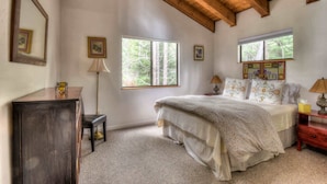 A bedroom with a wooden ceiling, two windows, a bed with floral bedding, a nightstand, a lamp, and a dresser.
