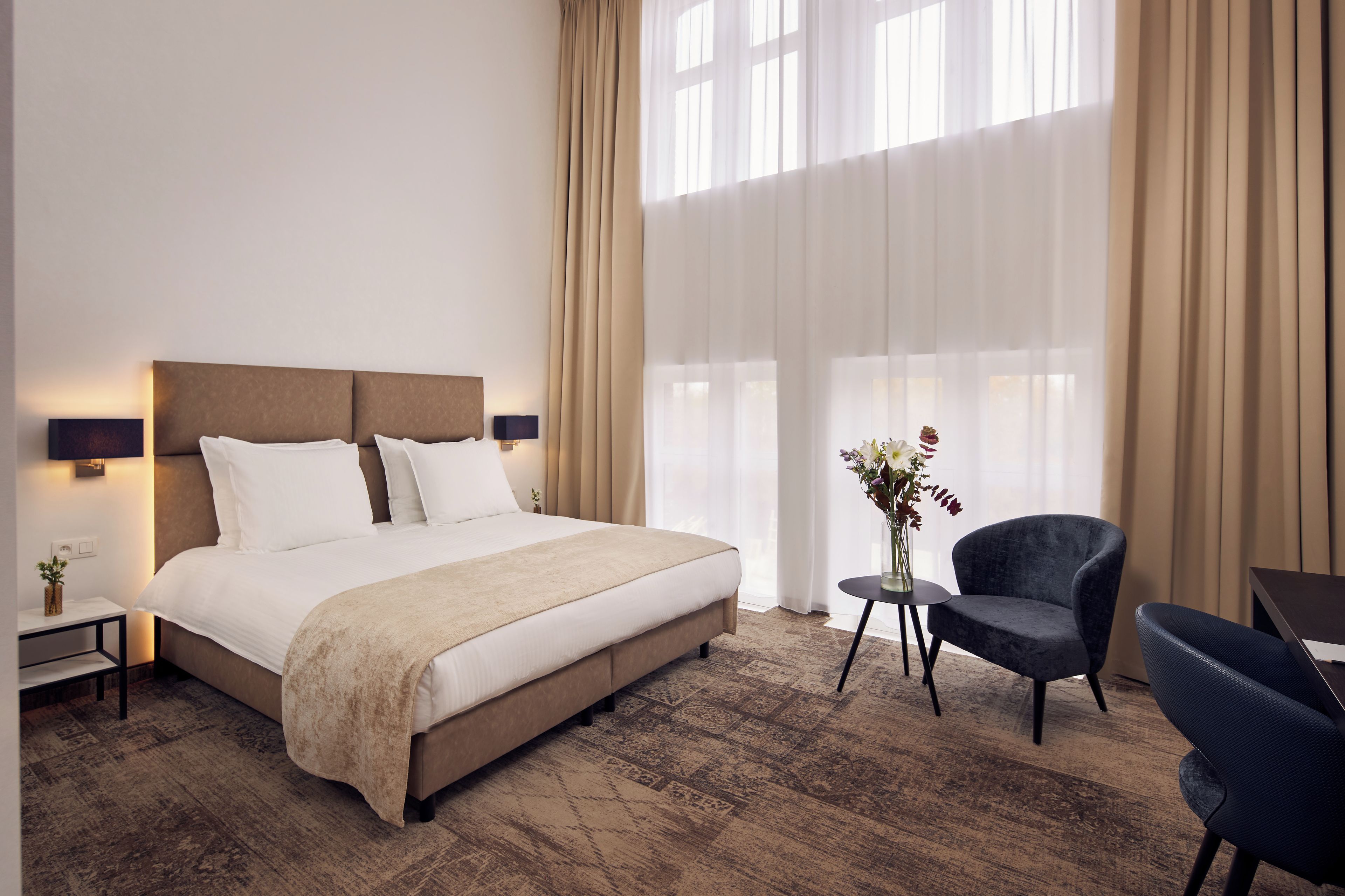 Hotelkamer met een groot bed, een klein tafeltje met een lamp, een stoel en uitzicht op het landschap door dunne gordijnen.