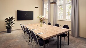 Uma sala de conferências com uma mesa comprida, cadeiras, uma planta em vaso, um vaso com flores, uma TV de tela plana e janelas grandes com cortinas.