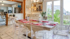 A modern kitchen with white cabinets, a dining area with clear chairs, and a large window offering a view of the outdoors. - Phil'blue by Interhome (Saint Philibert)