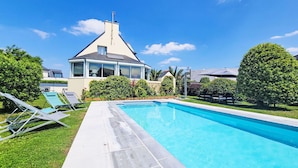 A swimming pool with a tiled deck, a house with a large window, and a well-maintained lawn. - Phil'blue by Interhome (Saint Philibert)