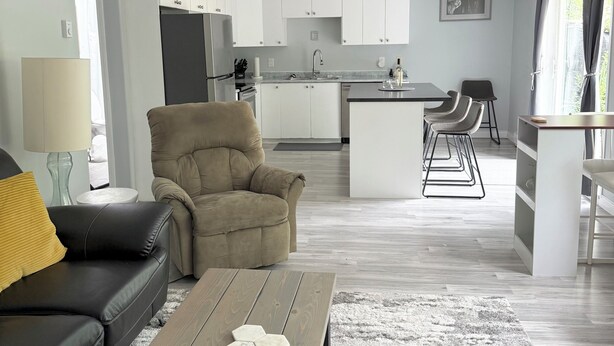 En moderne stue med en sofa, et sofabord og en beige lænestol. I baggrunden ses et køkken med hvide skabe og et spiseområde med barstole.