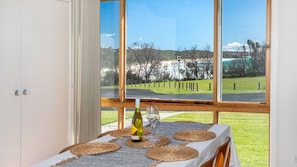 A dining area with a table set for two, a bottle of wine, and a glass on a table with a view of a grassy landscape and trees through large windows. - FREE NIGHT. Pay for 2 stay for 3 (Dolphin Point)