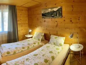 A room with two beds, each with a nightstand and a lamp. There is a window with curtains, a painting on the wall, and wooden paneling.