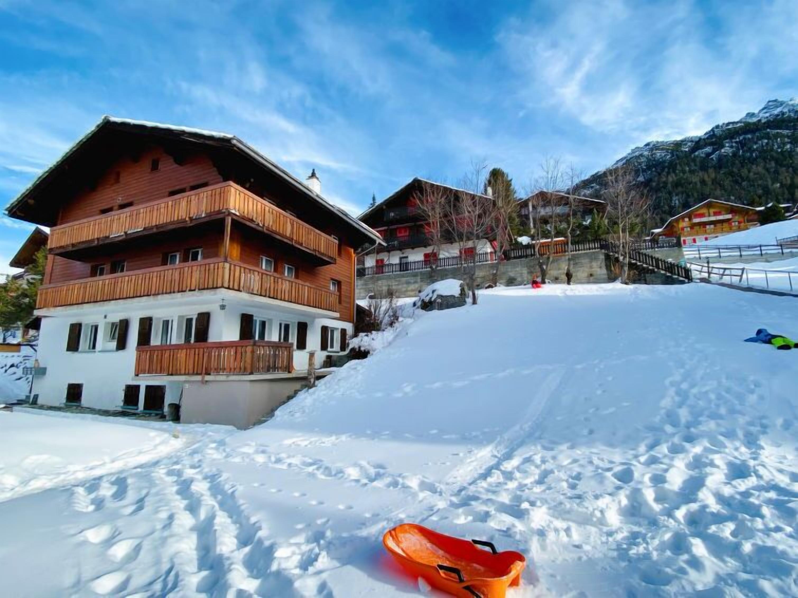 A multi-story wooden chalet with balconies, set against a snowy mountain backdrop.