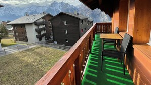 Ein Balkon mit Holzgeländer, Tisch und zwei Stühlen. Im Hintergrund sind Gebäude und schneebedeckte Berge zu sehen.