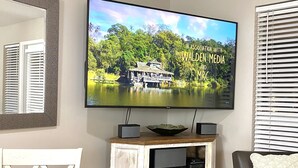 A living room with a television mounted on the wall displaying a scenic image, a wooden dining table with a decorative bowl, and a comfortable seating area with a chair and a sofa. - The Turtles Escape (Destin)