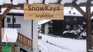 Een houten bord met de tekst 'SnowKaya Jungtalblick' boven een overdekt terras met houten balustrade en een trap die naar beneden leidt.