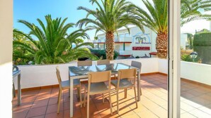 A balcony with a glass table and chairs, palm trees, and a building with a sign in the background. - El Gato I by Interhome (Llançà)