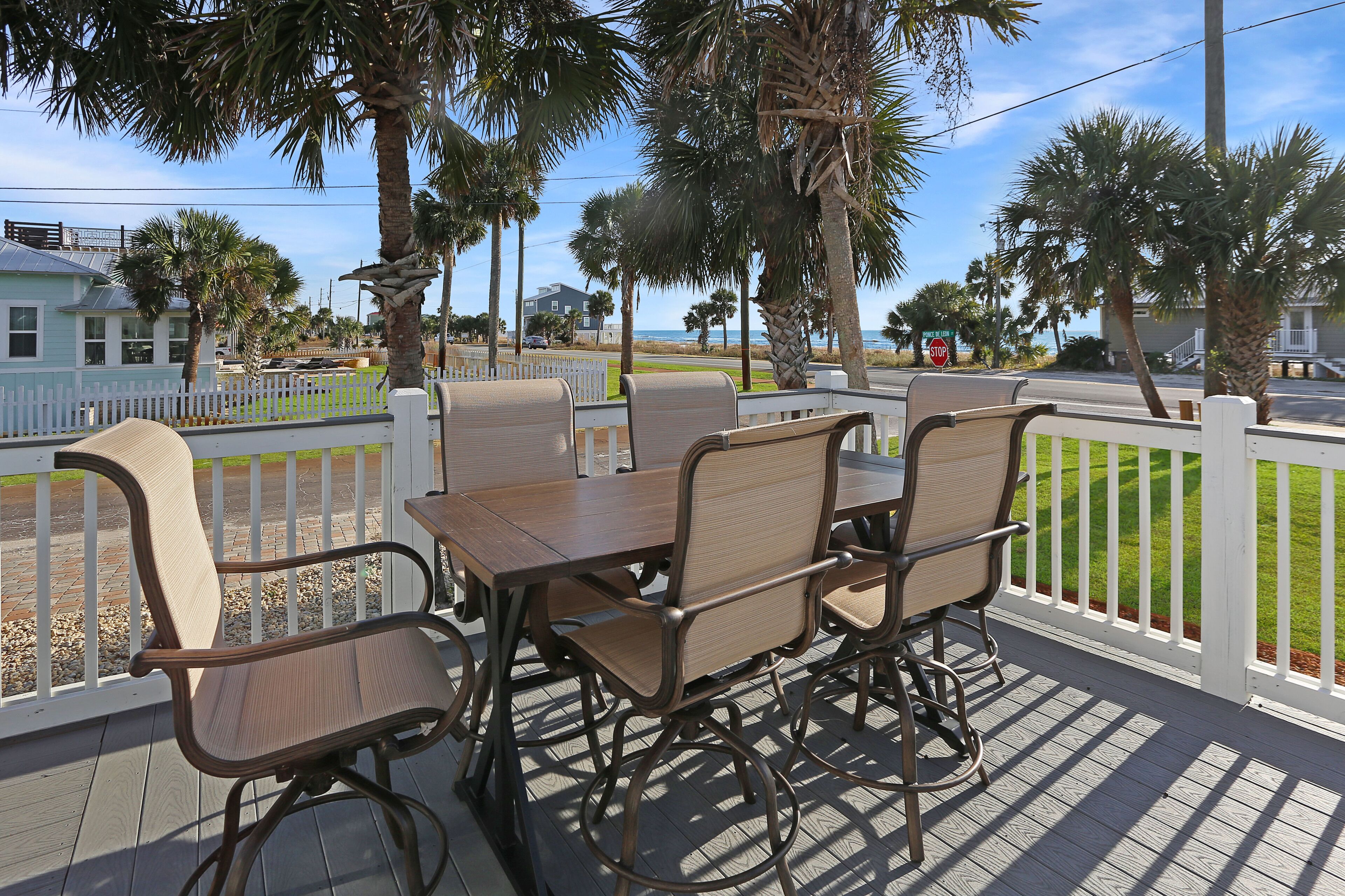 A wooden deck with a table and chairs, surrounded by a white fence, overlooking a beach with palm trees and houses.