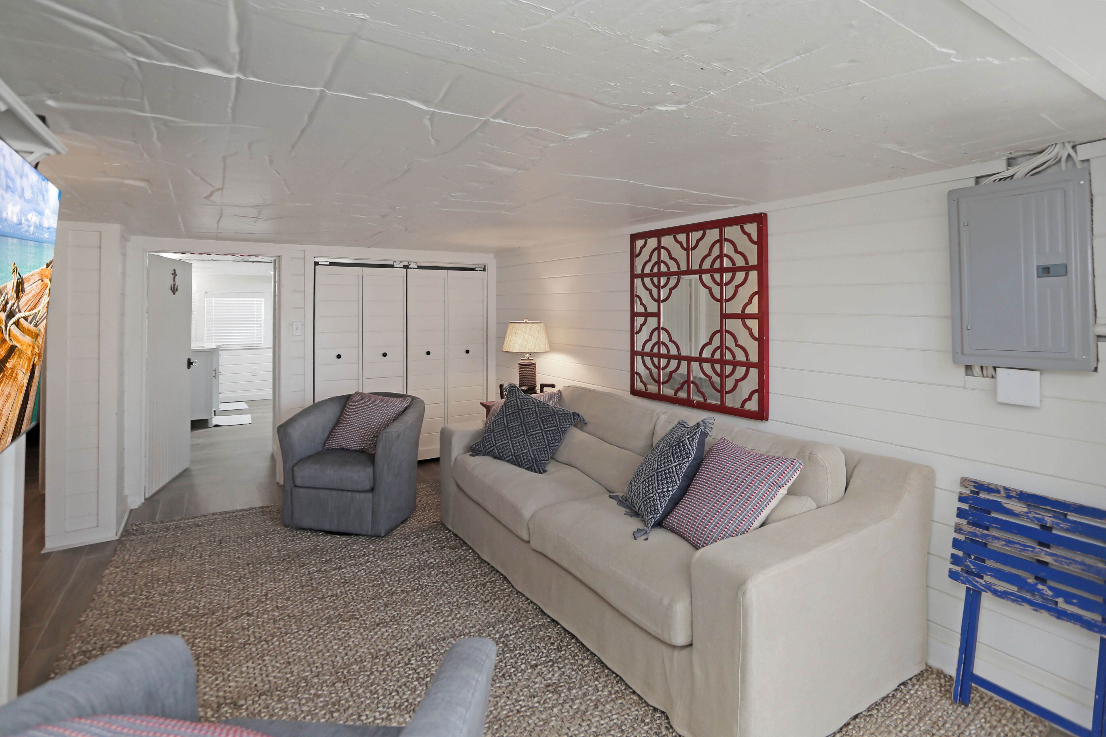 A living room with a sofa, armchair, and a lamp. There is a door leading to another room and a wall-mounted electrical box.