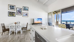 Una cocina moderna con una isla grande, armarios blancos y una zona de comedor con sillas de mimbre. Hay fotos enmarcadas en la pared y un televisor en la isla.