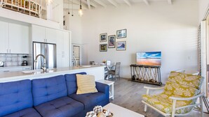 Un espacio moderno para estar con un sofá azul, una butaca amarilla con estampado, una mesa de centro blanca y una cocina con armarios blancos y electrodomésticos de acero inoxidable.