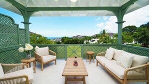 Un espacio al aire libre cubierto con mobiliario de mimbre, una mesa de madera y vistas al océano y casas al fondo.