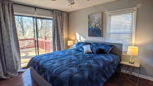 A bedroom with a large bed, a ceiling fan, a window with blinds, and a view of a deck.