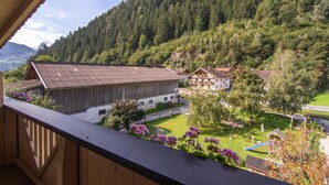Un balcon donnant sur un village rural, avec ses bâtiments traditionnels, une aire de jeux et une colline boisée.
