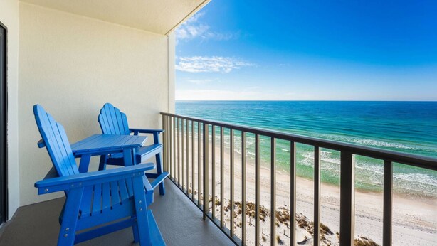 A balcony with a table and two chairs, overlooking a beach with clear blue water and a sandy shore. - Commodore 802 (Panama City Beach)