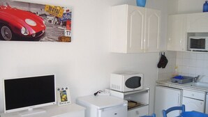A compact kitchen with white cabinets, a microwave, a small refrigerator, a television, and a blue chair.