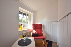 Une pièce d’angle avec une fenêtre, une chaise rouge, une petite table avec un vase et un radiateur.