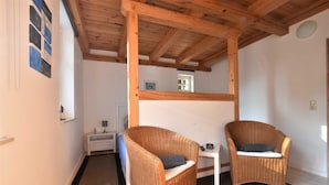 Una habitación con dos sillas de mimbre, una mesita blanca y techo de madera, con una puerta al fondo.